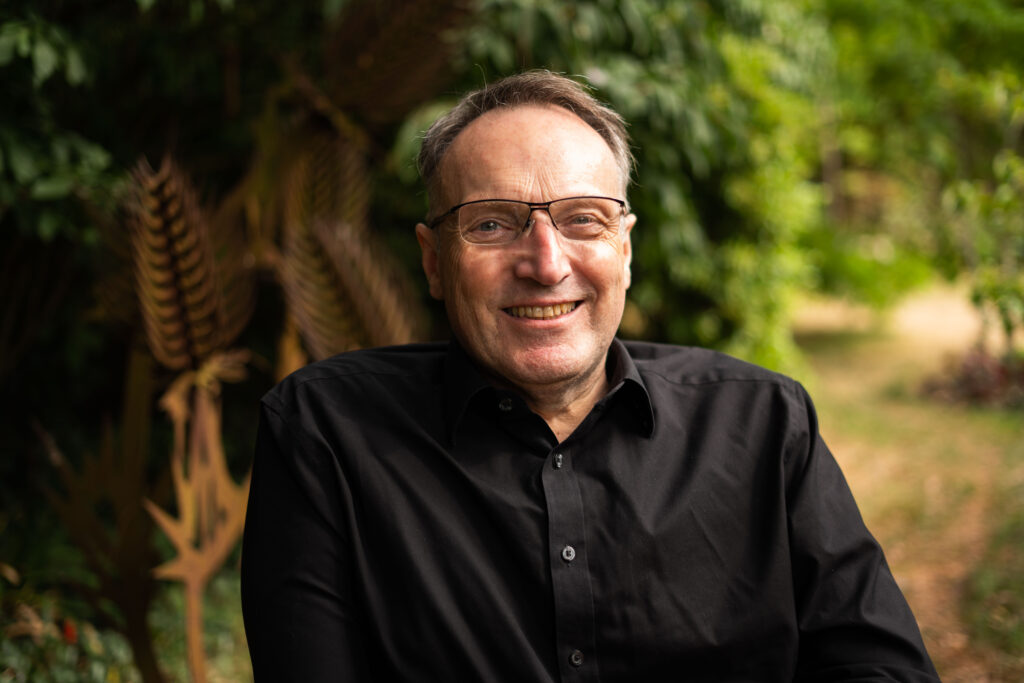 Photo of Andrew Miller, a white man wearing glasses. He is dressed in a black shirt and is smiling. He is sat in a wheelchair outside, there are out-of-focus plants in the background.