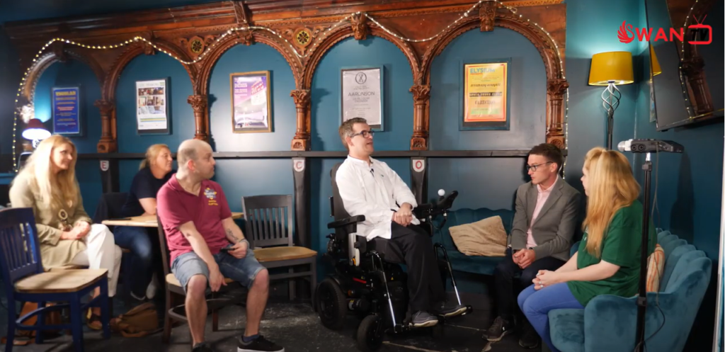 An image from on set. Lee is sat in the centre in his chair and is facing a man and woman sat on the blue sofa in front of him. Three people sit behind Lee on wooden chairs, watching as an audience.