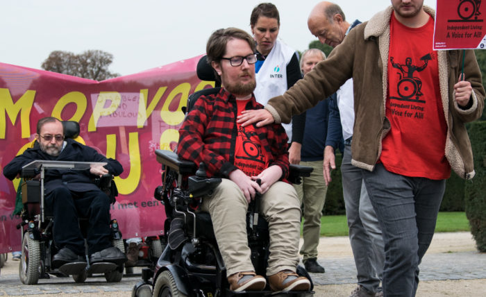 Miro Griffiths is driving a power wheelchair, with a personal assistant supporting him by holding his chest. The assistant is holding a placard about disability rights.