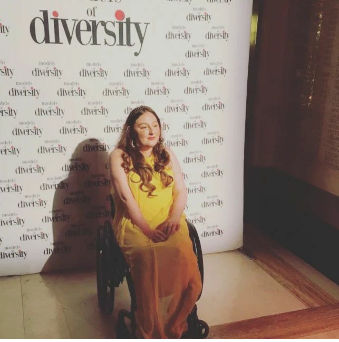 Dark haired women in a wheelchair wearing a yellow dress, in front of the models of diversity poster.