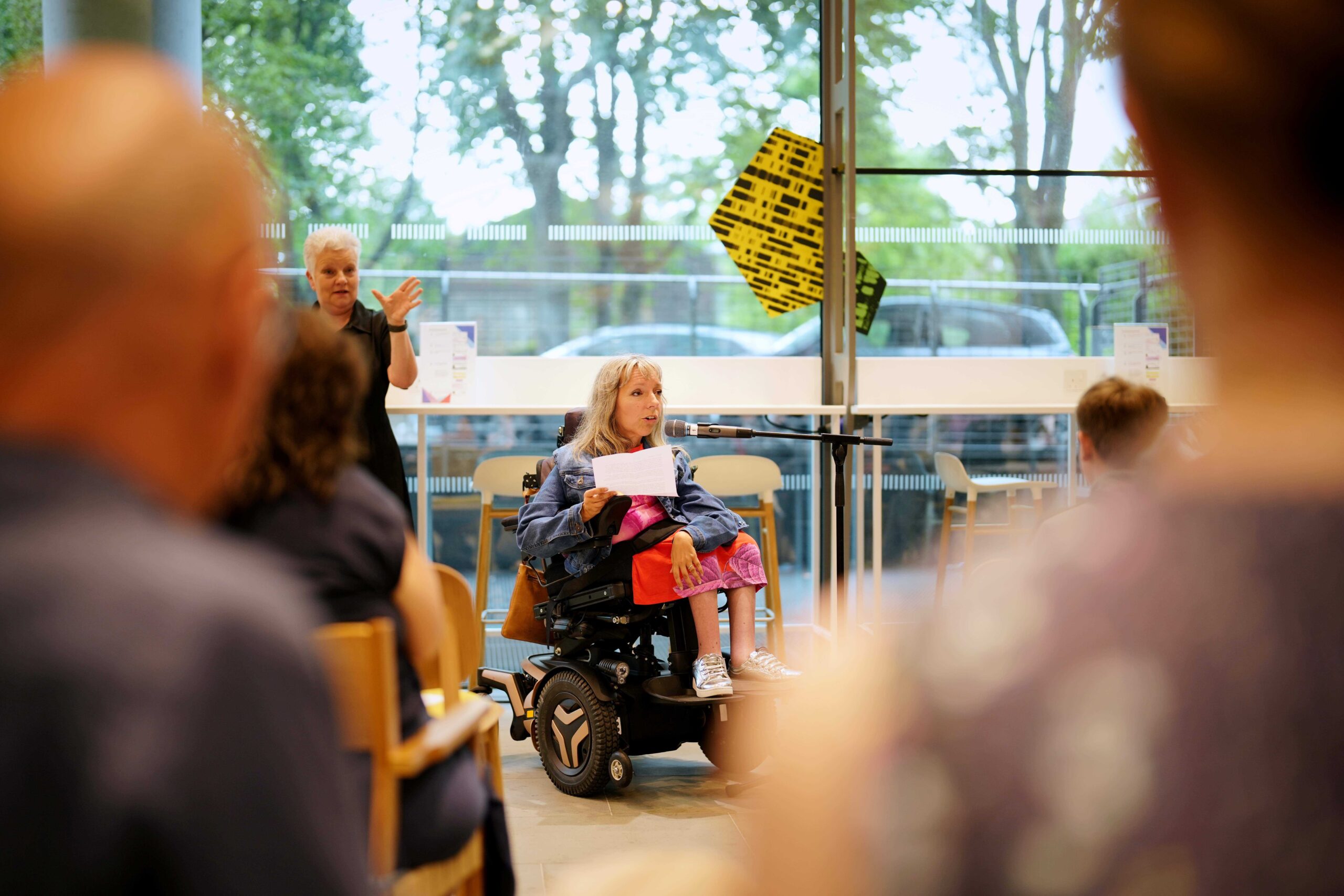 ows of people seated with their backs to the camera and facing towards Esther who is making a speech seated in her wheelchair. There is a large window behind her. Esther is a white woman in her late 40â€™s with mid-length blonde-grey hair, wearing a denim jacket over the top of a pink and orange t-shirt dress with silver shoes.