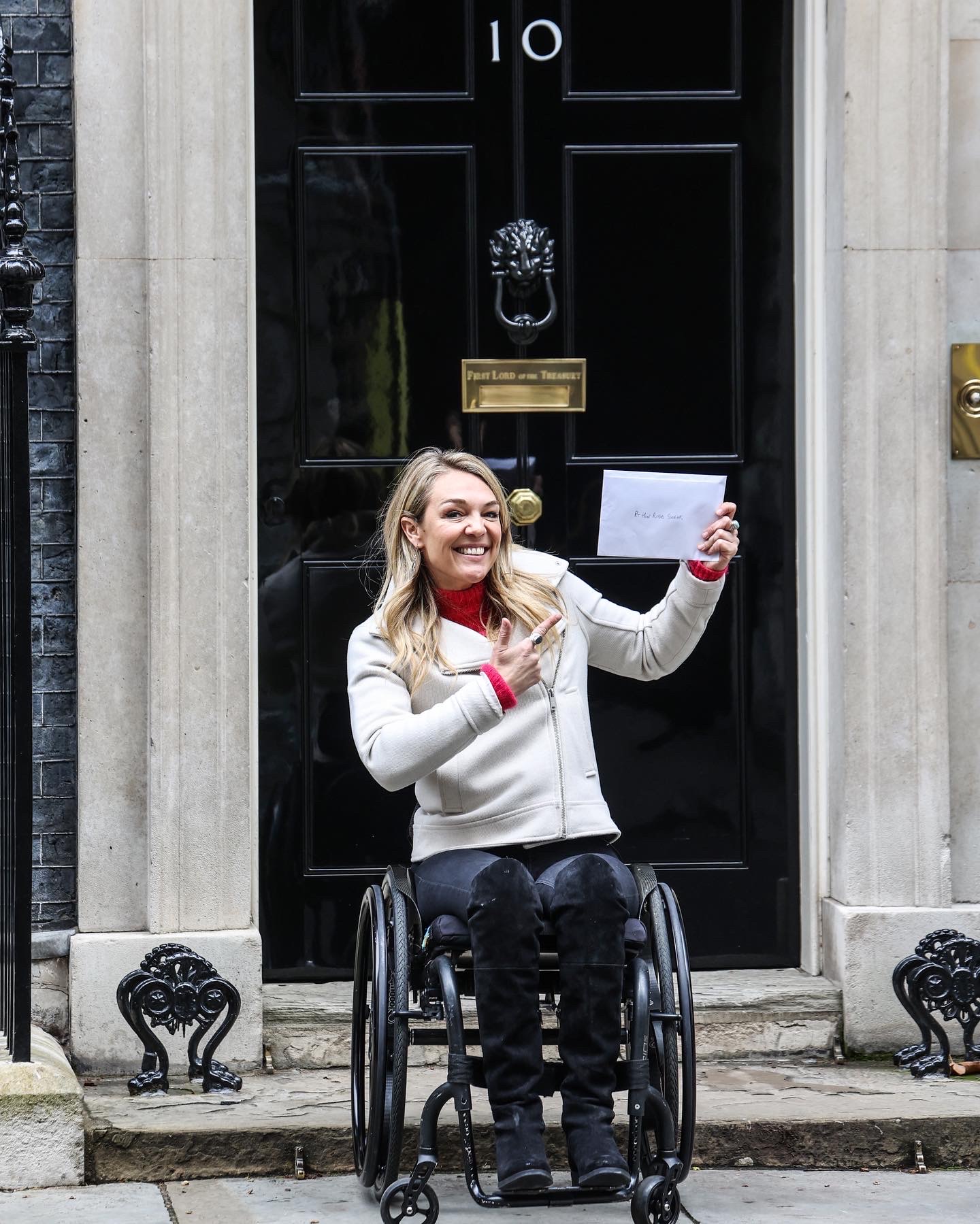 I'm a blonde woman in a manual wheelchair, sat on the steps on 10 Downing street holding an envelope and smiling.