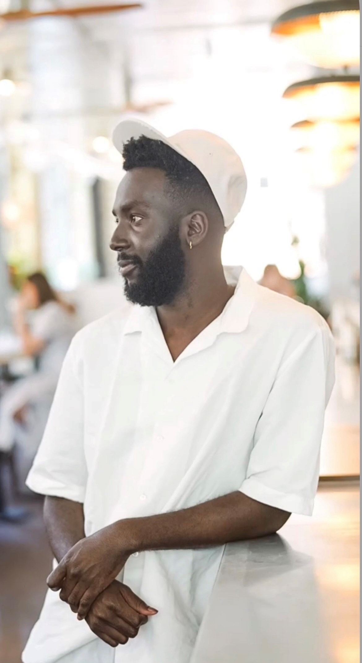Chris is a young black man, dressed in a crisp white polo shirt with a v-necked collar. Chris has a beard and wears a white cap, which is tilted backwards slightly, to reveal some of his hair. He also wears an earring on his left ear. 