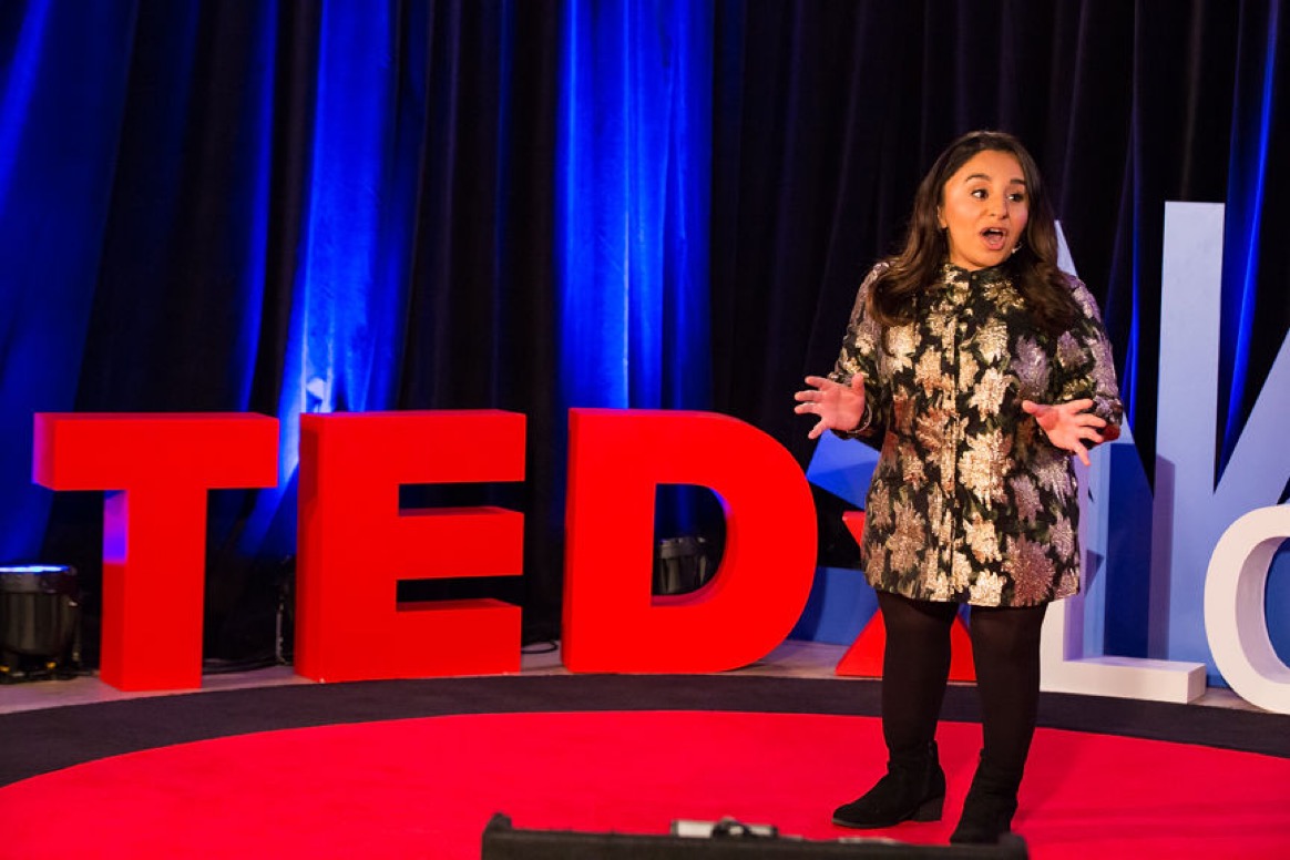 Full length photo of a South Asian disabled woman, Shani Dhanda, delivering a TED X talk on stage.