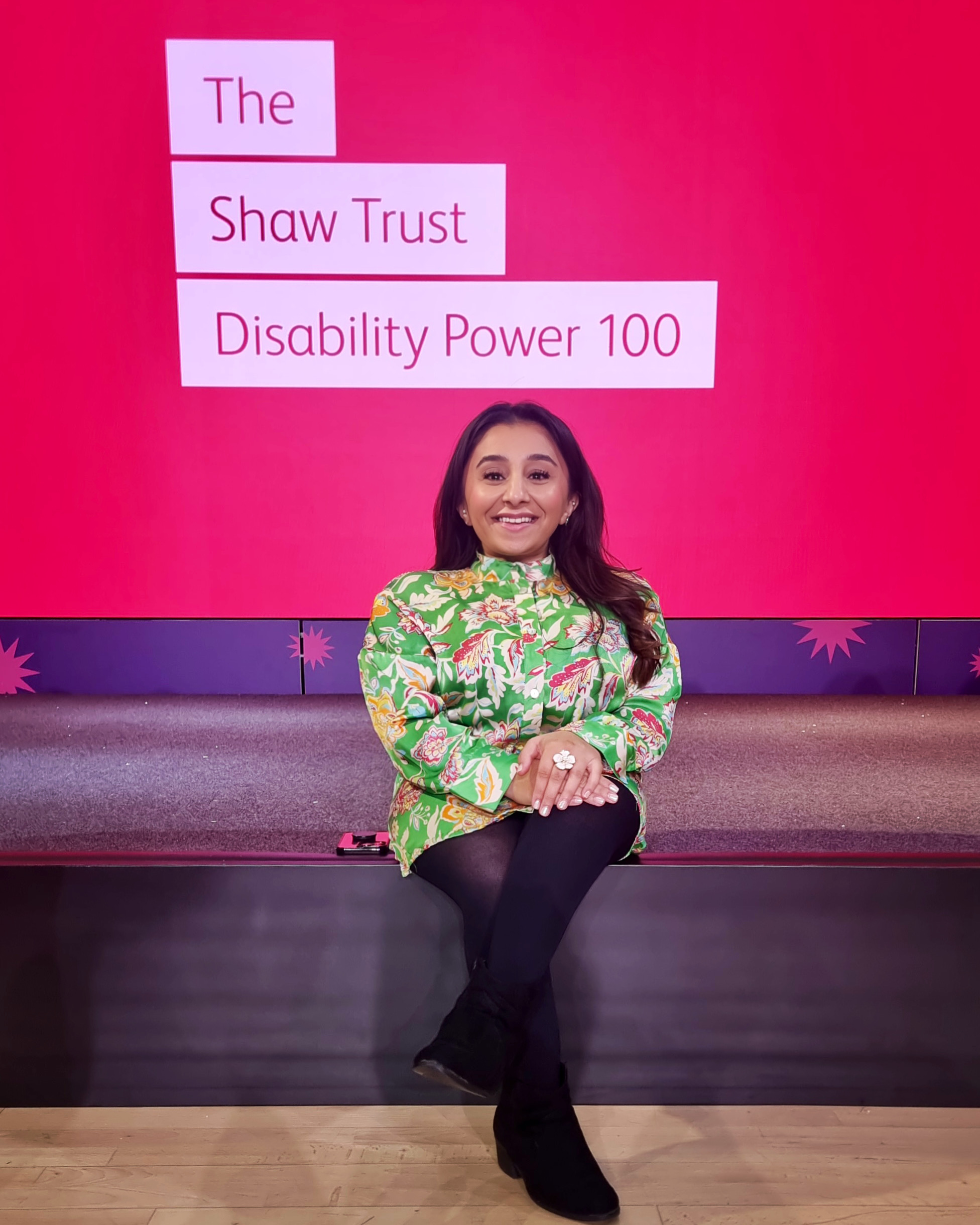 An image of a woman seated on the edge of a stage with dark hair over her shoulder, and a screen behind her displaying the Shaw Trust Disability Power 100 logo