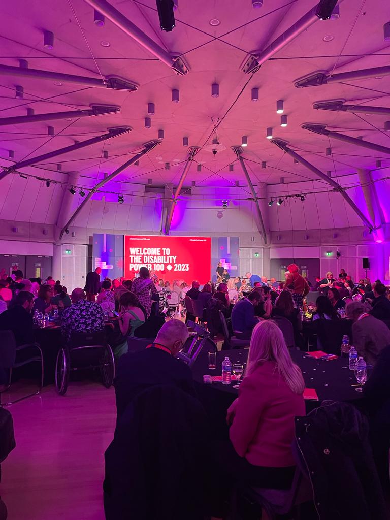 A photo from the back of the event room. There are pink and purple lights and people sat at the back around tables and in the front on rows of chairs. The screen behind the stage is red with white text "Welcome to the Disability Power 100 2023."