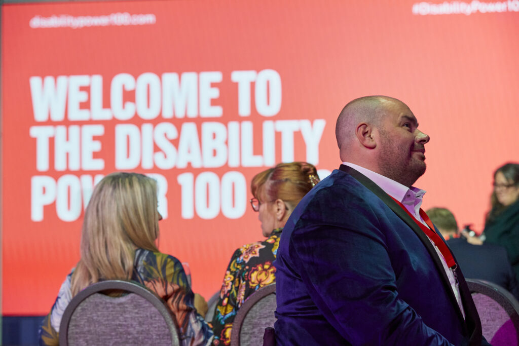 Text on a screen reads "Welcome to the disability power 100£. A man in smart clothes is talking to someone off screen