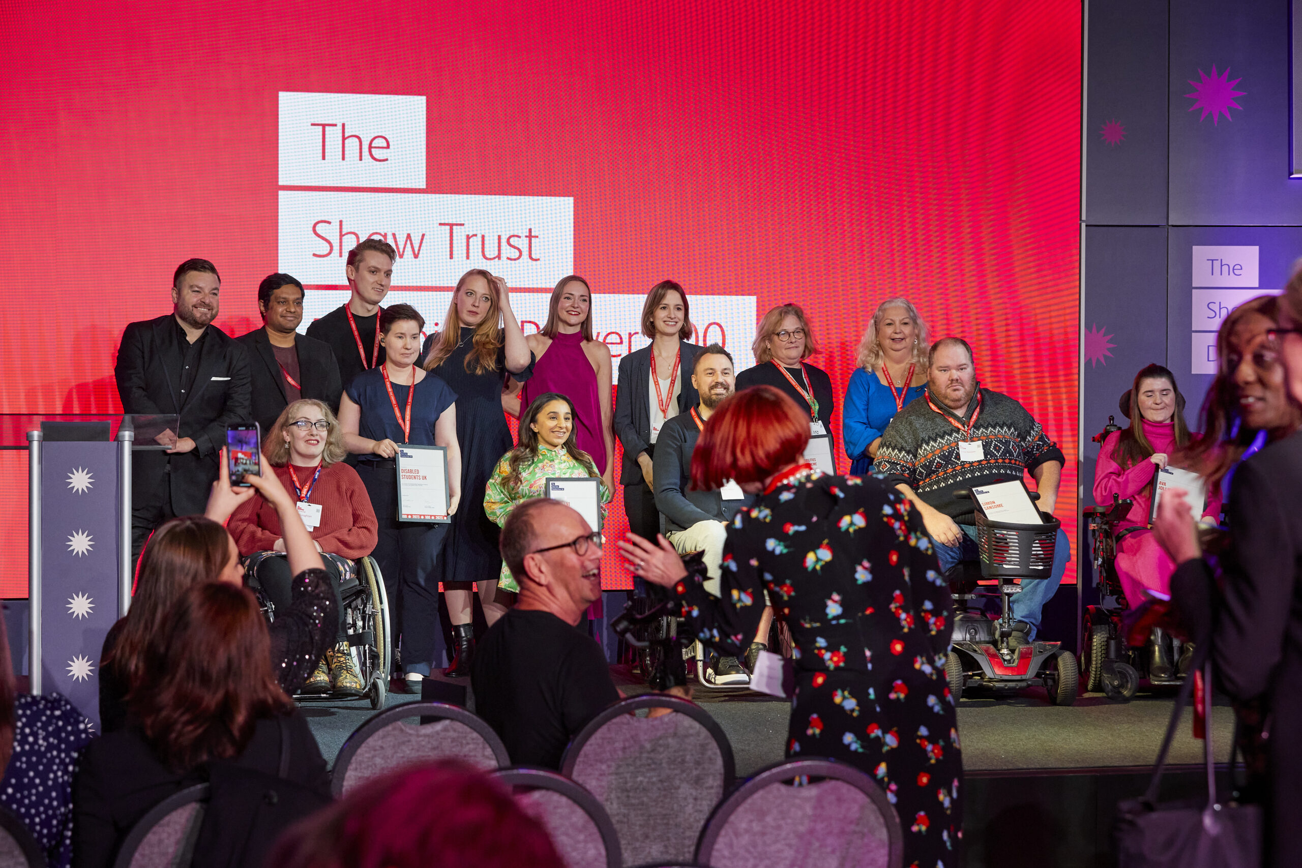 photograph of the winners from 2023 on a stage as a group, they are looking slightly to the right and are in front of a red screen with white writing saying The Shaw Trust Disability Power 100