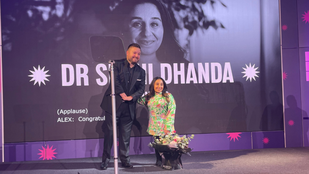 Shani, an asian woman with short stature is stood behind a big bunch of flowers and next to Alex Brooker, who is wearing a blue suit. The screen behind has a black and white image of Shani and her name written in white text.