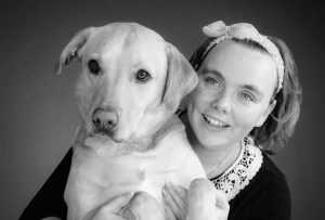 Chloe (a short-haired lady) hugs a Labrador assistance dog and smiles for a photo. Black and white image