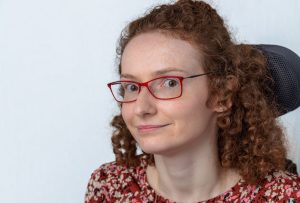 A head and shoulders shot of a white woman with aburn curly hair and red glasses. She has a brown, rust, white and pink patterned top on and gold necklace. There is a headrest behind her head and the background is white.