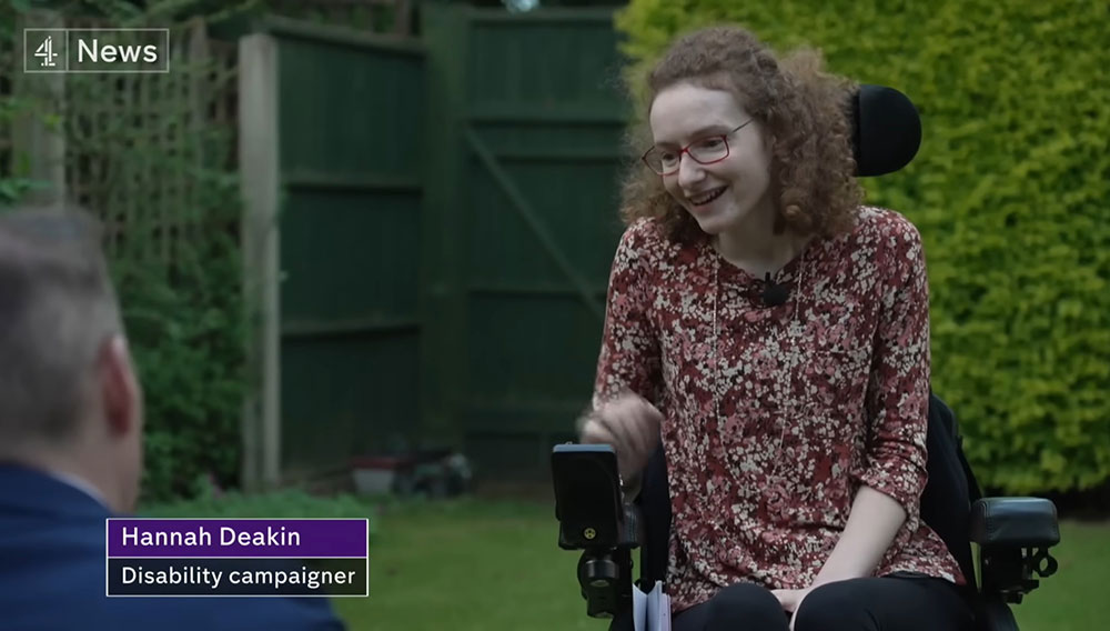 Hannah, a white young woman sitting in her powerchair in her garden speaking to a newsreporter. In the bottom left hand side of the photo the words 'Hannah Deakin Disability Campaigner' in white writing with a Purple background.