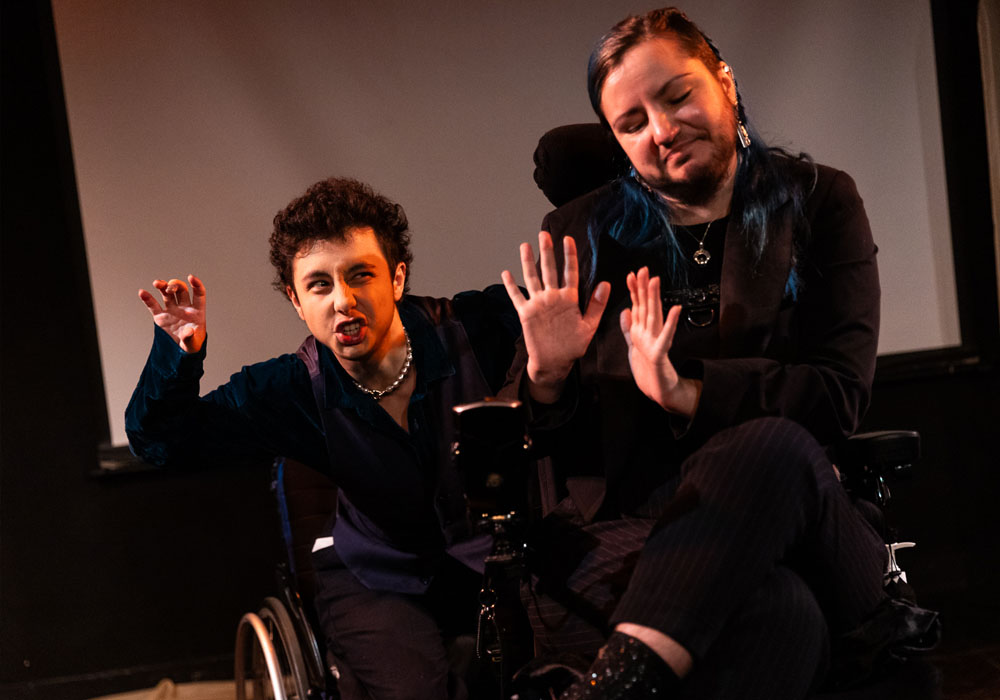 Performers Sam Zelaya, and Max Marchewicz performing on stage as part of The Crip Monologues. Sam, a mixed-race young man with light skin, dark curly hair, brown eyes and a slim build in a manual wheelchair, lurks both menacingly and comedically behind Max, a white person, with long blue hair, a dark beard, who is sat in their powered wheelchair, making a face as if they're not bothered by Sam.