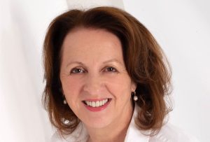 Jacqueline Winstanley, a woman with shoulder-length brown hair, is smiling warmly. She is wearing a white blouse with a collar, and pearl earrings. The background is a simple, light-colored setting, highlighting her friendly expression.