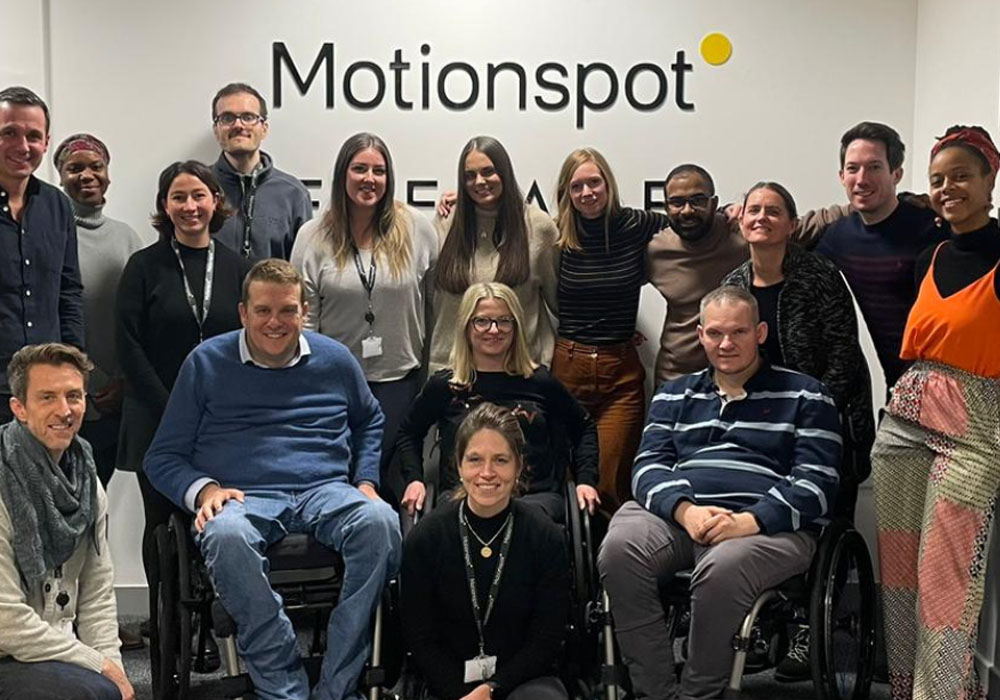 16 members of Motionspot's diverse team, smiling together in front of the Motionspot logo in their design studio