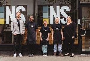 A group of 3 men and 2 women standing in front of a building with large windows and the letters "NS" displayed. The three central people are wearing "NS" aprons. They are all smiling and standing in a row, creating a friendly and welcoming appearance.