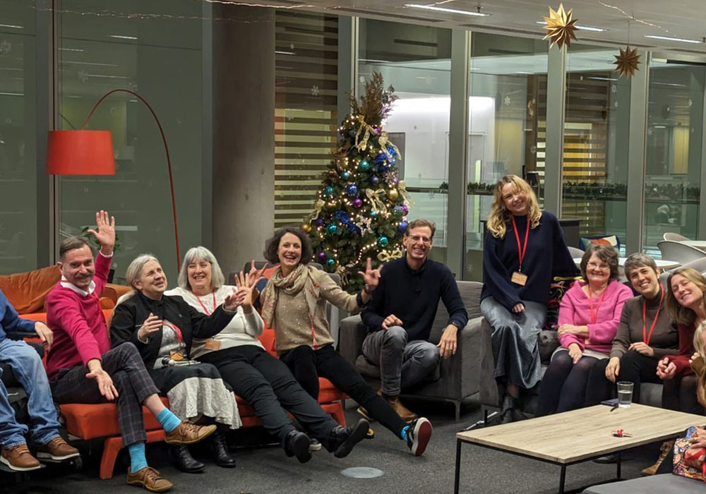 Open's UK team at the office in London with a Christmas tree. 3 men and 10 women of differing ages sitting on sofas in a U-shape. Some are wheelchair or scooter users, one uses a cane, one is blind and one is a BSL user, a number are neurodivergent, have chronic or mental health conditions, but of course a sighted viewer can't see all of that.