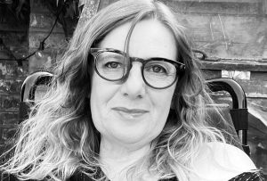 A black and white headshot of a middle-aged white woman with long curly hair, sitting in an electric wheelchair. She is wearing round glasses and smiling. She is in her garden.