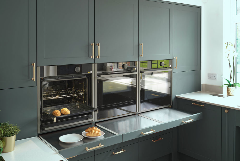 A bank of kitchen cabinets in dark green with 'hide and slide' ovens and pull-out shelves underneath them.