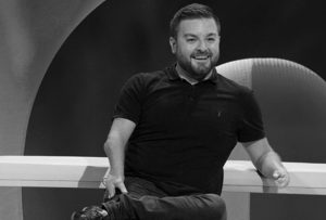 An image of alex brooker, he wears a dark polo neck, and is sitting on the set of the last leg.