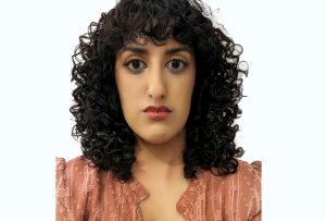 A Brown woman with shoulder-length curly black hair, a left-side nose ring and two lip piercings, wearing a peachy-pink top in front of a white background.