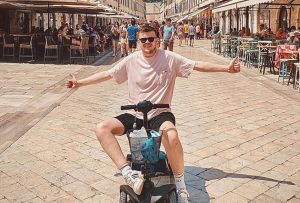 Angus is sitting on a mobility scooter, facing the camera with his arms spread wide and giving two thumbs up. He is wearing a light pink t-shirt, blue denim shorts, white Adidas trainers, and sunglasses. The photo is taken on the main street of Dubrovnik, Croatia. Cafes with outdoor tables line both sides of the street, and the light-coloured Mediterranean-style buildings frame the scene. In the background, an old church tower with a domed roof is visible.