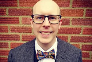 A man wearing glasses, a paisley bow tie (navy background with a motif of gold and orange), a white shirt, a navy waistcoat, and a grey/blue blazer jacket in front of a red brick wall.