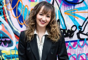 A white woman smiling with dark brown curly hair, wearing a black blazer standing in front of a bright street art backdrop.