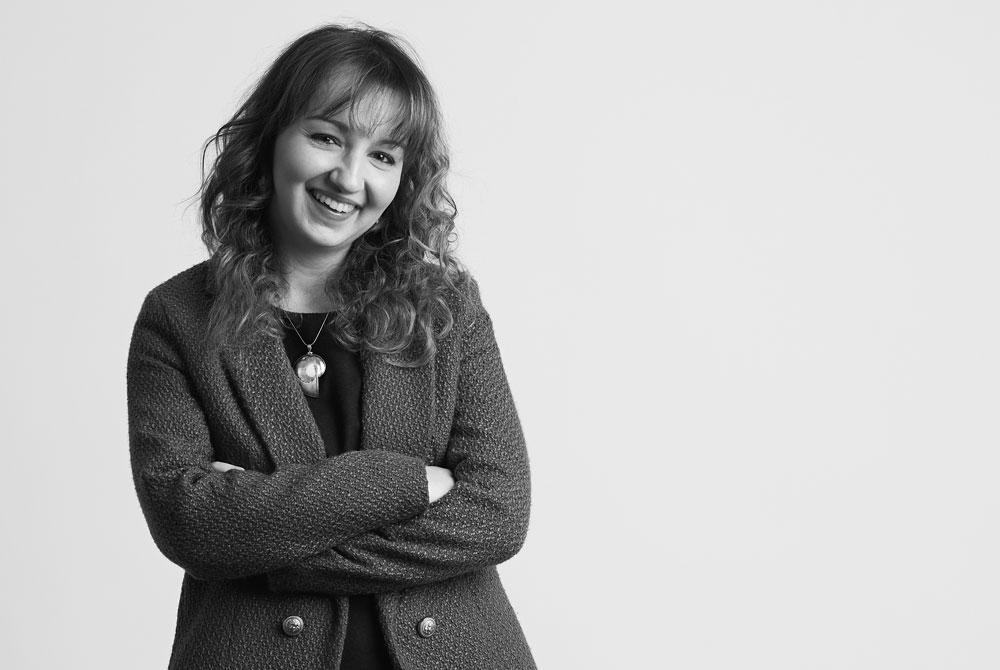 "A white woman smiling with dark brown curly hair, wearing a black blazer standing in front of a bright street art backdrop.