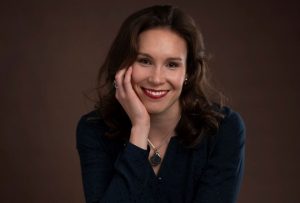 A picture of Beth, a young lady, wearing business dress and sitting on a stool leaning towards the camera and smiling