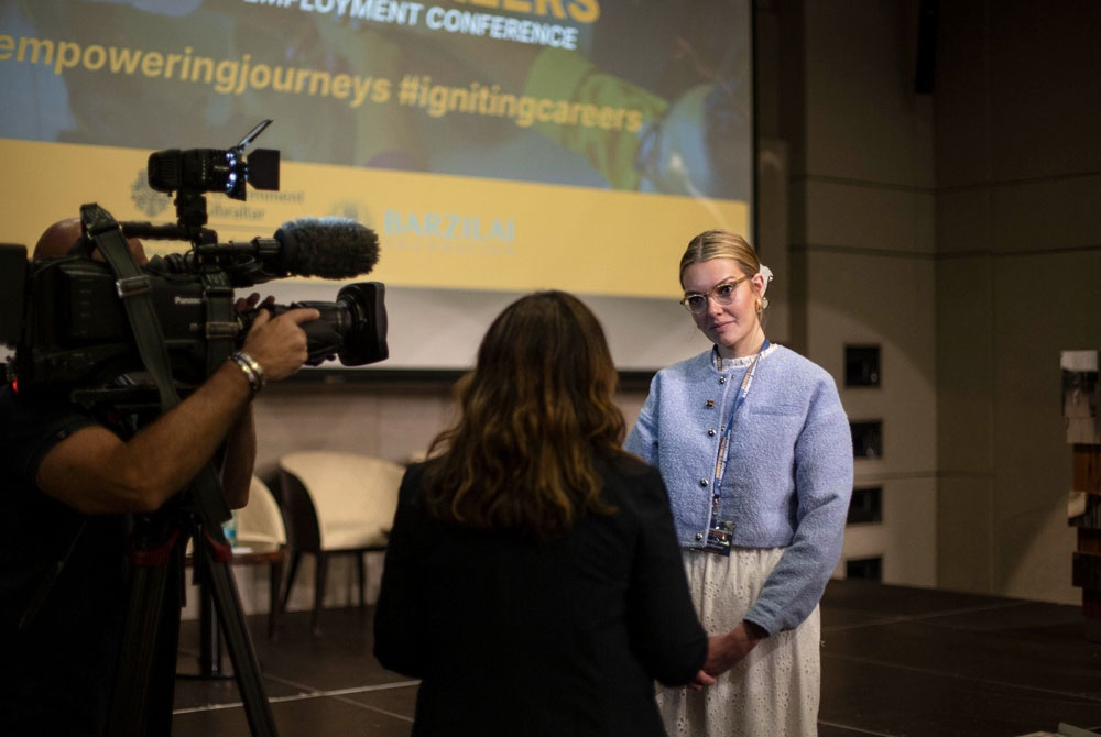 Image shows Celia a white female with blonde hair tied back off her face talking to a woman with a micrphone, there is a cerma facing her. Behind her is a stage and a lectern where she has just finished giving a keynote speech.