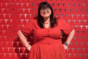 Chella wears a red dress and red acrylic necklace laser cut into the shape of the word ‘period’. She poses in front of a wall covered in wallpaper featuring a print of silver uteruses on a red background. Chella has long, straightened dark brown hair with a fringe and wears large black framed glasses. Image Credit Photo taken with permission at the Lauft (Flow) Exhibition at the MEK Museum of European Culture, Berlin