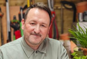 Man with dark hair in a green top, inside a potting shed with tools in the background and plants.