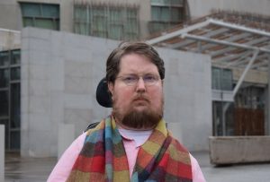 Image of Miro Griffiths, with short brown hair, beard, and round rimmed glasses staring at the camera. He is wearing a pink shirt and a multicoloured scarf is draped across shoulders. His wheelchair headrest is visible and he is outside in front of a concrete building.