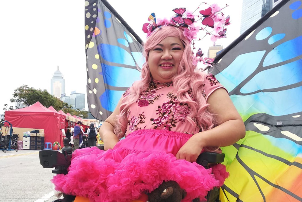 a woman in a power wheelchair with pink curly long hair with little butterflies as decoration. She had a huge colourful butterfly wings behind her back. She wears a pink qipao and a tutu dress. She has a rainbow leggings and leather boots. She places her hands on her waist to signifies the confidence of wonder woman.