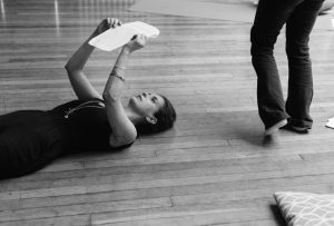 Black & white image of a woman dressed in a black jumpsuit lying on a wooden floor on her back holding up pieces of paper to read them. She has dark hair, bracelets, and a long necklace. Her skin tone is white passing. Also in the image there is the lower half of another person walking, wearing black trousers and white socks.