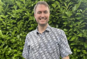 tall white middle-aged man smiling, standing in front of green shrubbery wearing a batik shirt with a diagonal pale-blue-and-black pattern