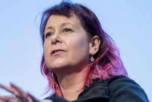 A brown and pink haired woman looking out as though she is about to speak.