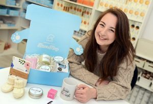 A dark haired white woman stood smiling beside an open blue gift box.