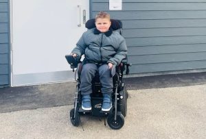 Image shows 11 year old Fraser in his powered wheelchair outside the Changing Places facility in his local park, that he had campaigned for, and had the honour of opening. He is wearing a grey coat with a fur collar, tracksuit bottoms and trainers. His hair is gelled and he is smiling at the camera.