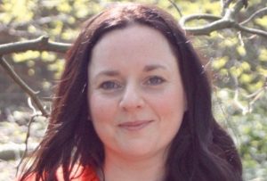 A white female with long brown wavy hair, wearing g a red jumper.pers standing in front of trees.