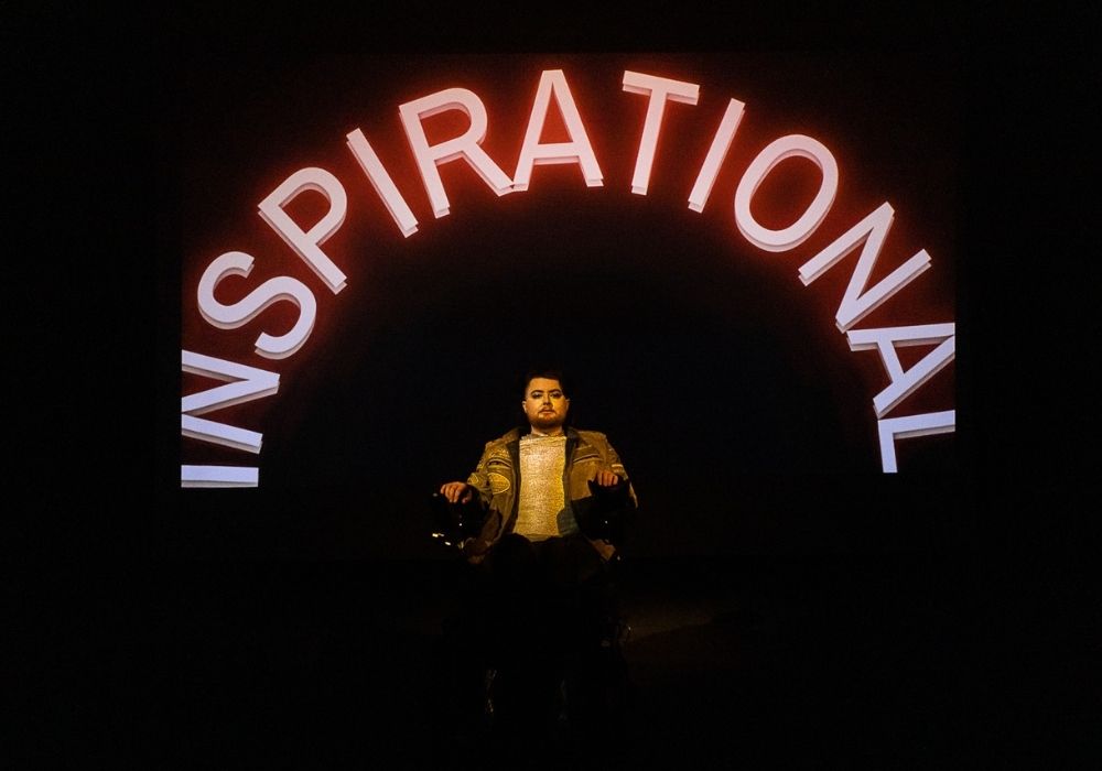 "A white person in an electric wheelchair, under a large “INSPIRATIONAL” in reddish neon-style lighting./ They are wearing a leather jacket and silver shirt. Image credit: Shona Louise"