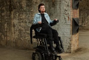 A white person in an electric wheelchair, lifted to standing height. They are wearing a blue denim jacket and have dark red hair and a dark red beard. Behind them is a brick wall.