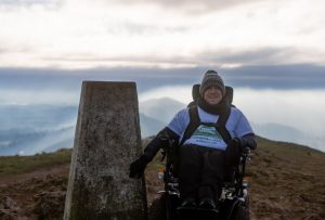 Josh is touching a stone on the top of a sunny but cold and misty mountain in his black all-terrain wheelchair.