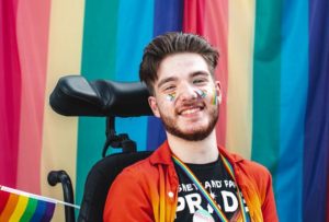 Lee with dark hair, beard against a pride background wearing rainbow stickers.