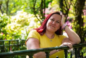 Maddy has red hair, she is wearing a yellow tshirt and leaning on a railing surrounded by greenery.