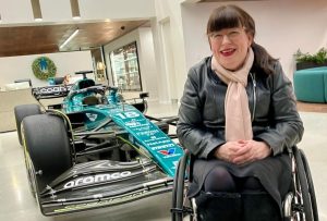 Mary, a white middle-aged woman with dark hair and glasses, wearing a black outfit and pale scarf. Mary is a wheelchair user and behind her is a green Formula 1 car on display.