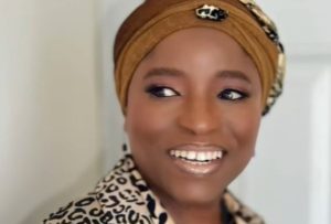 Funmi is a confident black woman with a warm smile, looking off into the distance. She is wearing a stylish brown flowery turban, a matching brown blazer, and a brown top. Her elegant attire complements her radiance, and her captivating smile lights up the scene.