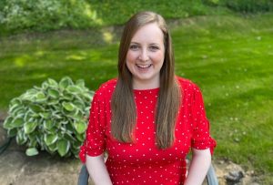 Natalie is a white woman with blue eyes and long dark blonde hair. She is wearing a red top with white polka dots. In the background there is grass and trees.