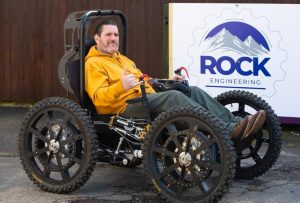 Nick is sat in the extreme all-terrain power mobility chair he used to ascend Yr Wyddfa in, called RockClimber, and is in front of a big sign saying Rock Engineering. He's wearing his signature Ability Over Disability orange hoodie, dark green jeans and usual cheeky chappy smile.